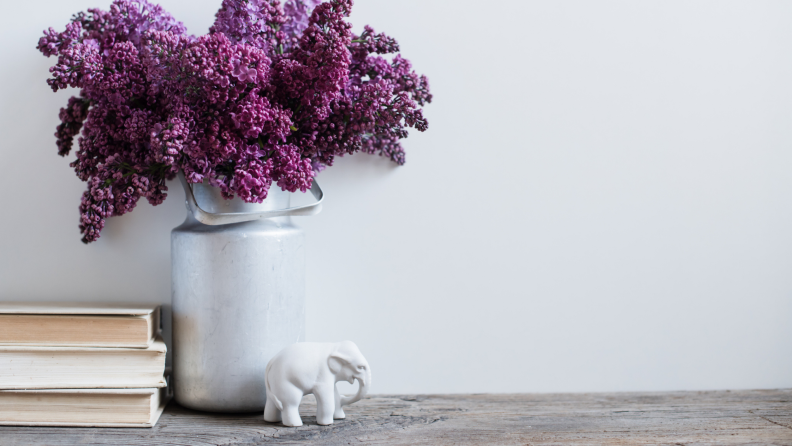 A stack of books, vase of purple flowers, and white elephant figurine sit on a shelf.