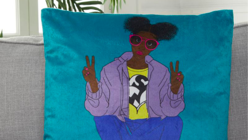 An image of a blue throw pillow with the image of a young Black woman with hair puffs and a lilac cardigan.