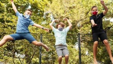 Three kids jump on a trampoline while wearing face masks.