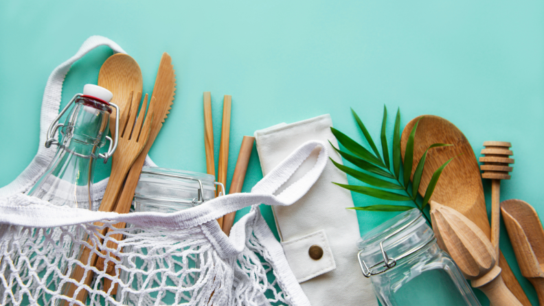 Cotton mesh bag, glass bottle, jar and bamboo cutlery on pastel green background.