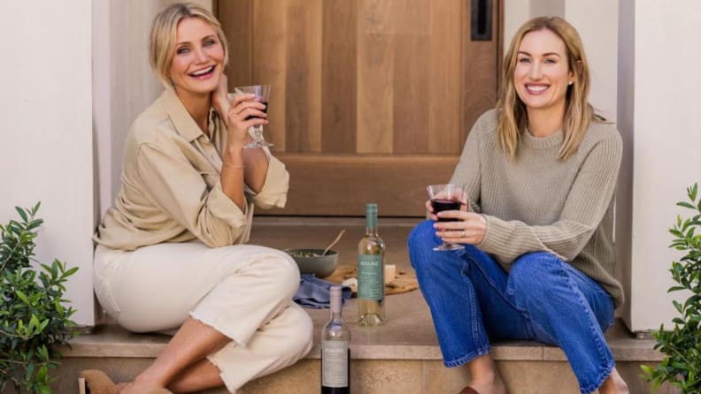 Cameron Diaz and Katherine Power posing on a stoop in front of a wood door and enjoying a glass of Avaline wine.