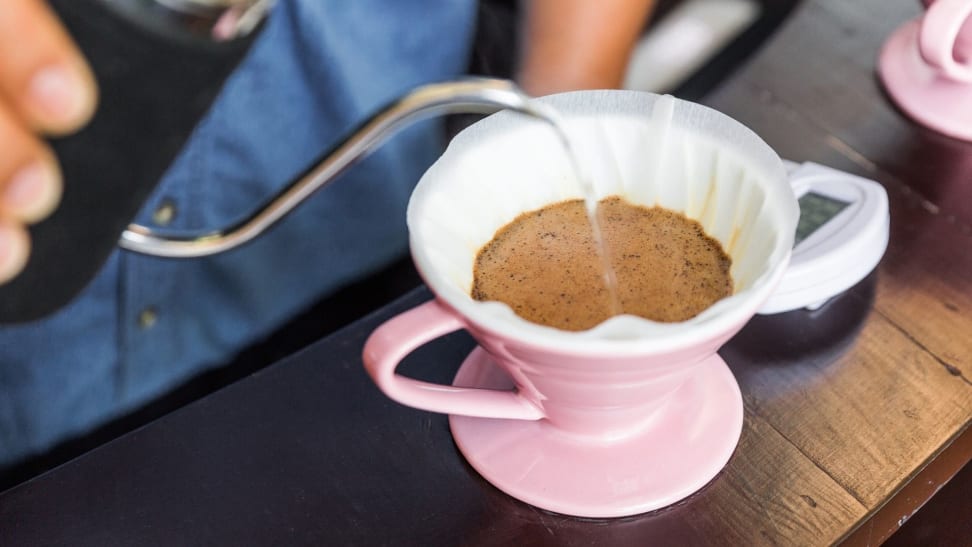 A person pours hot water into a pink pour-over coffee maker lined with a coffee filter and filled with coffee grounds.