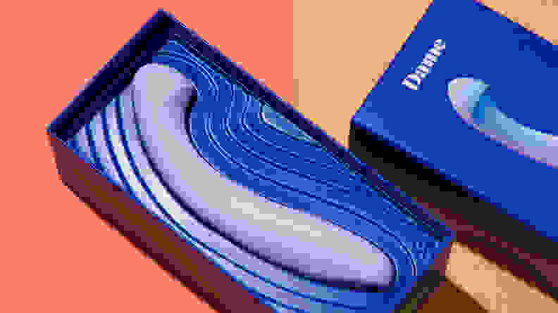 A blue Dame Arc vibrating dildo inside of its original box packaging on top of a two-tone orange surface.