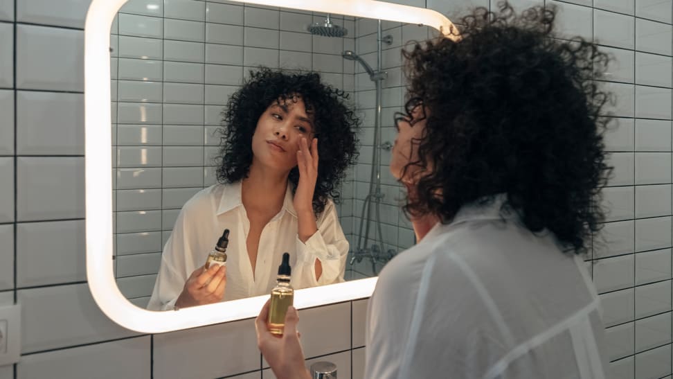 A person looking into a mirror touching their skin and holding a bottle of serum.