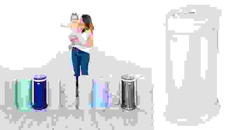 A woman holding a baby stands in front of a row of colorful Ubbi Steel Diaper Pails.