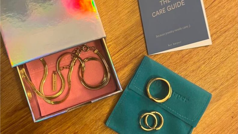 An image of a Hey Harper jewelry box in silver with a gold necklace nestled inside on a pink felt bag, accompanied by a blue felt bag on the outside with a gold ring and a pair of gold hoop earrings sitting on it. A Care Guide insert sits next to the jewelry.