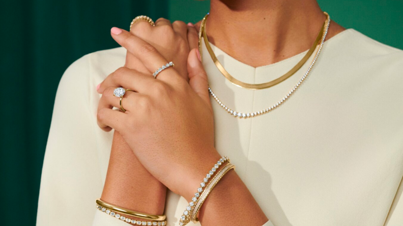 An image of a model wearing layered gold and diamond necklaces, diamond bracelets, and diamond rings on their hands.