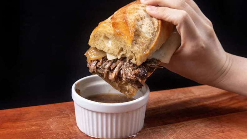 A hand holding a baguette sandwich with red meat, being dipped into a ramekin of sauce.