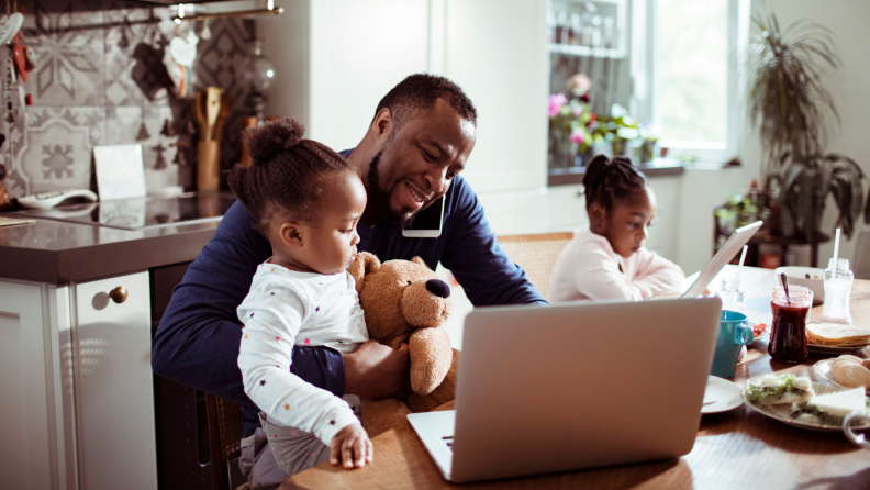 Father and two daughters in kitchen together as father is on talking on his cell phone and works on laptop computer.