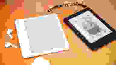 Two e-readers sitting on a wooden surface with a pair of glasses