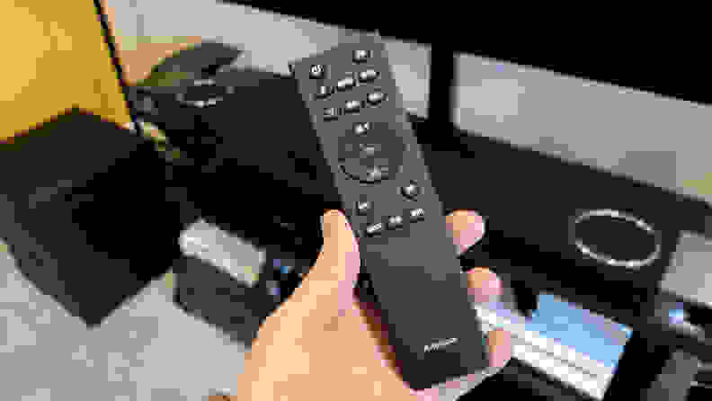 A black, rubberized remote control with white-lettered buttons is held before the black and silver soundbar on a black and brown console.