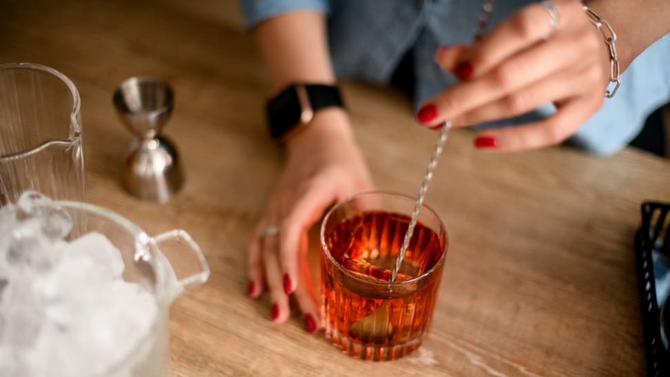 Woman mixing cocktail with bar spoon and jigger at kitchen counter