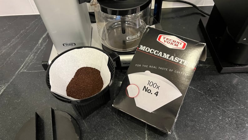 The box of No. 4 filters sitting next to brew basket full of ground coffee in front of the Moccamaster.