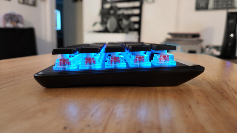 Side shot of the Roccat’s Vulcan II Mini Air keyboard with blue backlit display on top of wooden surface inside of room.