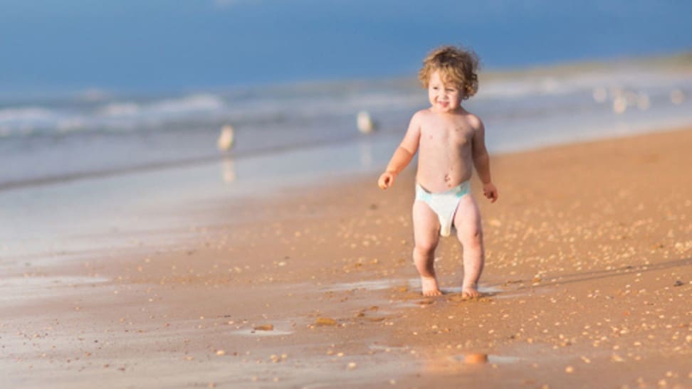 A baby in a diaper walking in the sand.