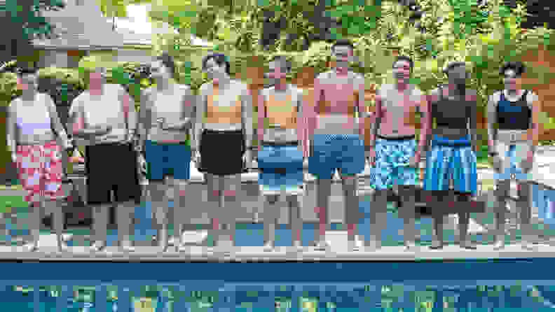 A bunch of people varying in size and shape standing poolside, all wearing binders and swim trunks, smiling and laughing.