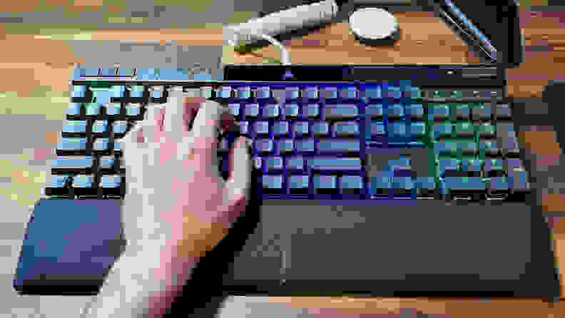 A left hand typing on a Corsair K70 Max RGB keyboard, with a wrist rest below it and full RGB on each key.