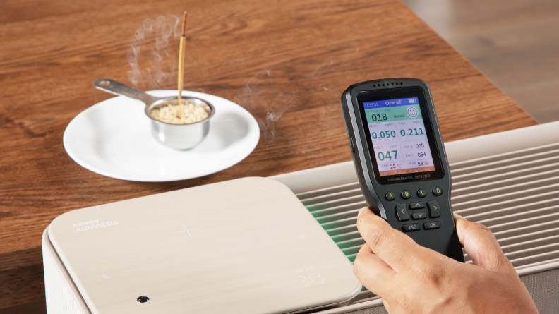 An incense stick burns on a small plate next to an air purifier, smoke rises, a hand holds an air quality monitor that displays data about the air.