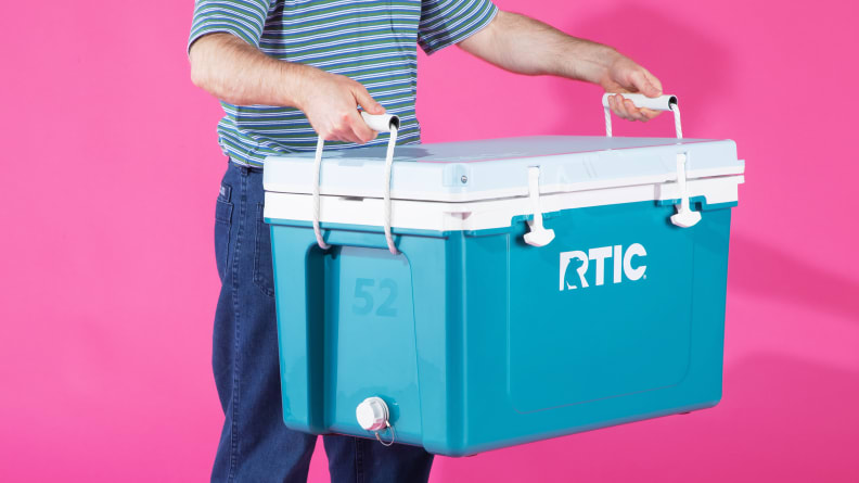 A man holds an Rtic cooler in front of a pink background.