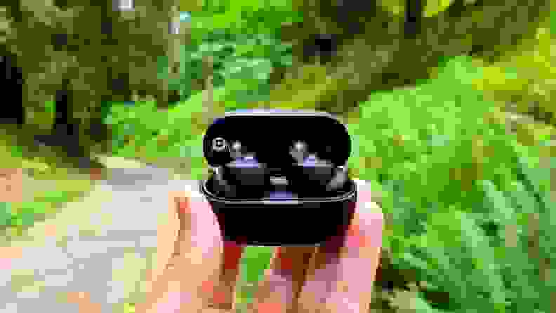 The black Sony WF-1000XM4 earbuds are shown in their case with a hand raised above lush green ferns and a mountain path