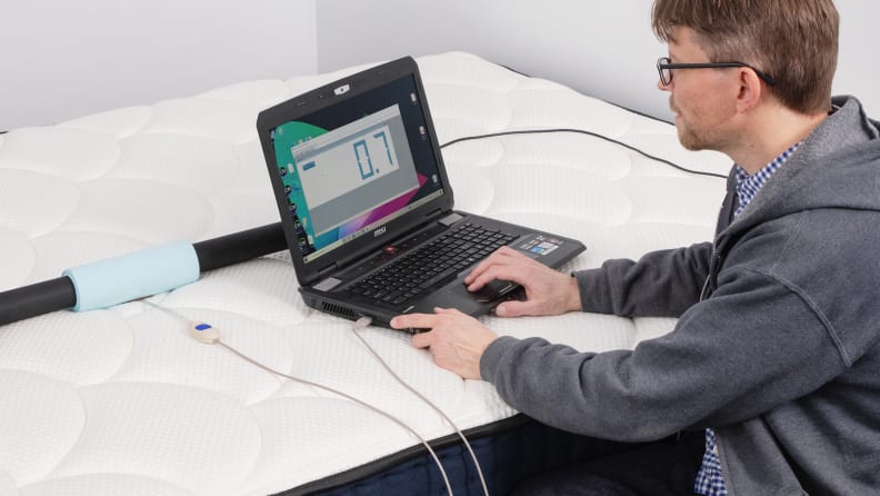 A person kneeling in front of a bed and looking at pressure point readings on a laptop screen.