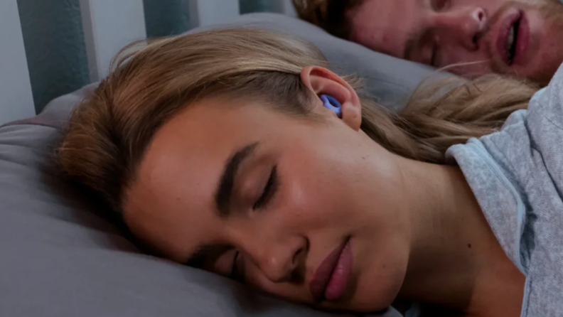 A woman sleeps with CURVD headphones in her ears while her partner seems to snore.
