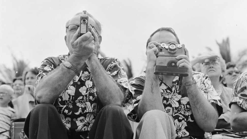 Two tourists in matching Hawaiian shirts poise with their cameras at the ready, awaiting the arrival of the Hula dancers on Honolulu's Waikiki Beach, in Hawaii.