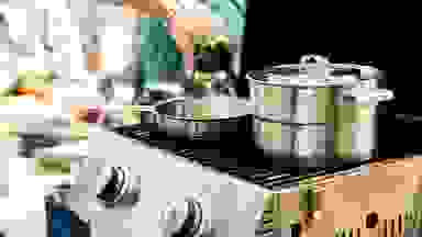 Stanley stainless-steel cookware is in use on a grill outdoors. Someone is pouring liquid from a bottle into a pan.
