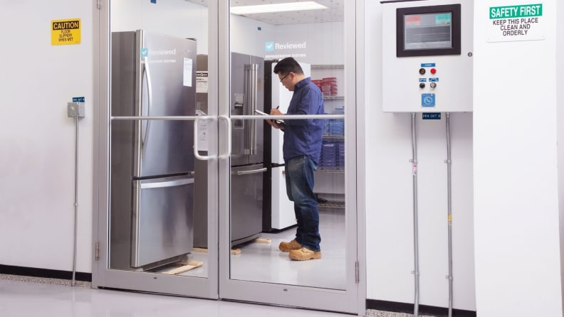 Lab technician standing in front of refrigerator inside of testing lab writes notes on clipboard.