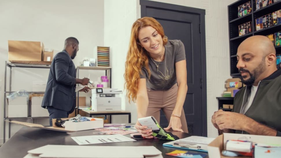 Woman holding patterned socks up for a coworker while another man uses the printer in the background