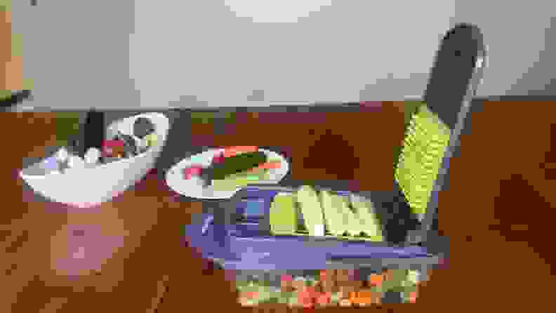 A bowl and small saucer plate filled with assorted vegetables next to the Mueller vegetable chopper with 3 ears of baby corn on top of the slicer.
