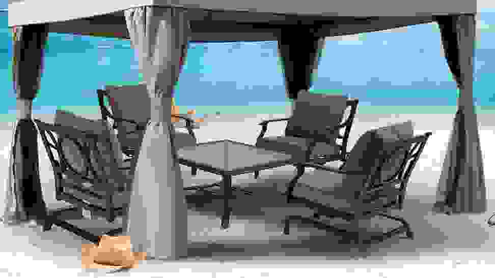 A gazebo tent gives shade to a table and chairs on the beach. The blue of the ocean is visible in the background.