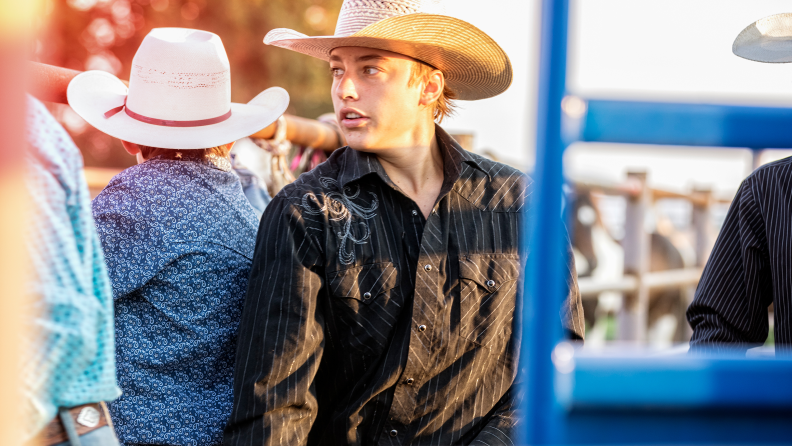 A man wearing a western shirt and cowboy hat stands in a crowd of other cowboys.