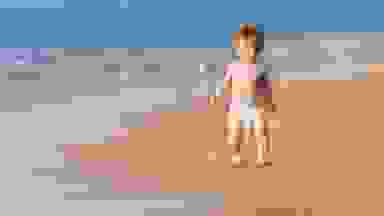 A baby in a diaper walking in the sand.