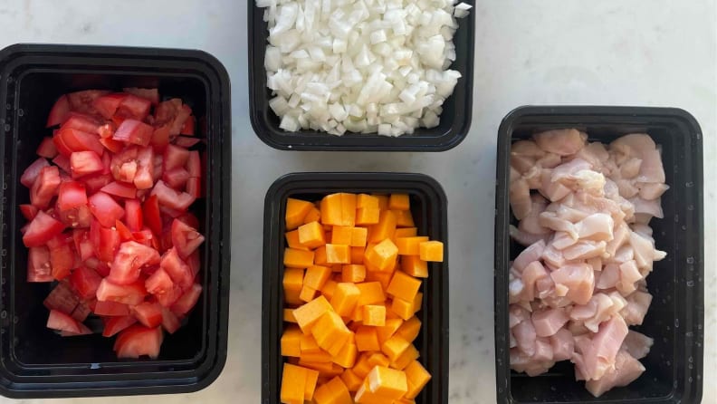 Four black plastic containers filled with diced tomato, sweet potato, onion, and raw chicken.