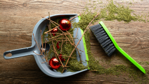 Pine needles, twigs, and red ornaments in a sweeping dustpan next to a handheld broom