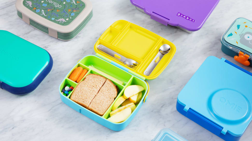Bento boxes, cheese stick, carrots, apple, a sandwich, and M&Ms in a lunchbox, laid out on a marble counter.