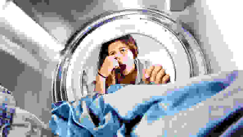 A shot from the back of a front-loader's drum shows a woman peering in at her laundry, pinching her nose in exaggerated disgust.