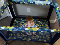 A small toddler sits on top of a patterned mattress inside of the Cosco Funsport Portable Compact Baby Playard.