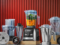 Several Vitamix blenders and accessories in front of a red backdrop