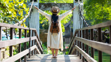 A woman in a beige outfit and hat stands on a wooden suspension bridge with her arms raised, surrounded by lush greenery.