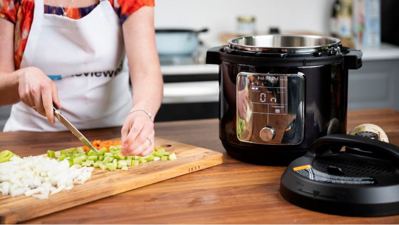 A woman wearing a Reviewed apron and chopping carrots, onion, and celery on a cutting board next to an Instant Pot. There is a blue pan on the stove behind her.