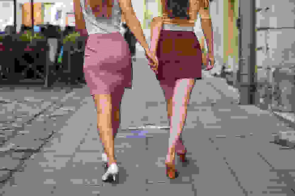 Two women walking, facing away from the camera. One wears a patterned pink mini skirt and the other wears a red patterned mini skirt.