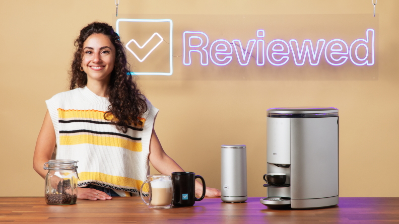 A person poses next to a table with a coffee maker, frother, and coffee cups.