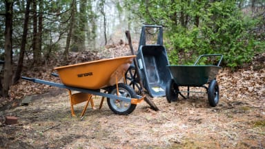 Three wheelbarrows—orange, black, and green—sit in a wooded area.