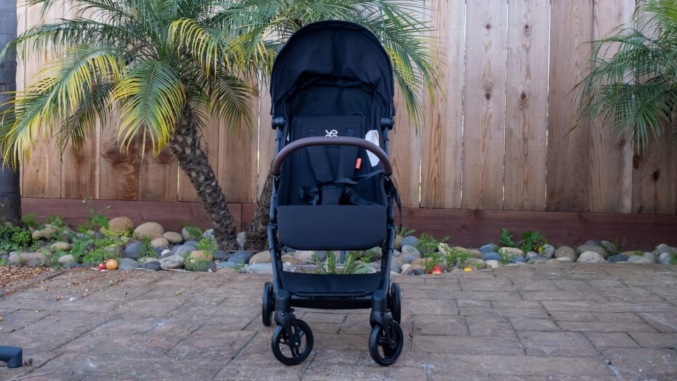 Black Mamazing stroller outside on a patio, set in front of a wooden fence and rock garden, with palm trees beside it.