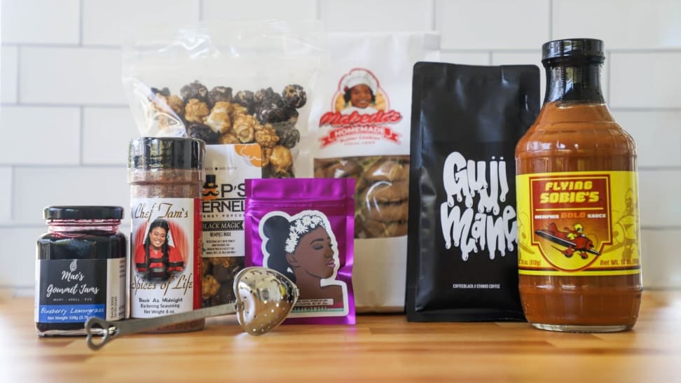 An assortment of spices and foods made by Black chefs are arranged on a light wood countertop with a white tile wall behind them.