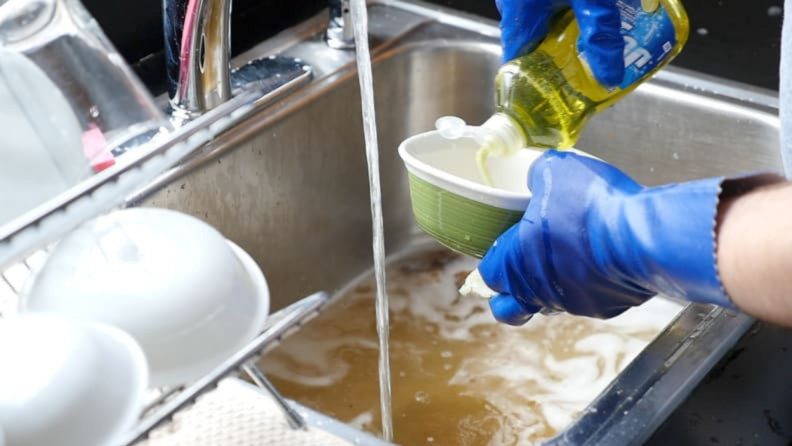 Hands wearing blue gloves washing dishes in a sink with soapy water.