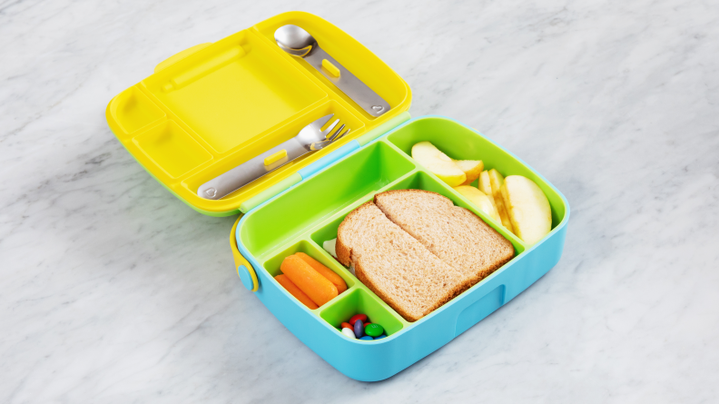 The yellow, green, and blue Munchkin kids lunchbox sits open on a marble counter with a sandwich, carrots, apple slices, and M&Ms in its compartments.