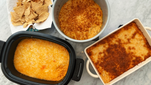 A bowl of tortilla chips and three pans of buffalo chicken dip laid out on a marble counter.